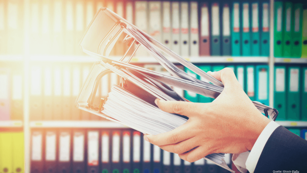 Eine Person im Anzug hält prall gefüllte Aktenordner im Arm vor einem Büroregal. Symbolbild für den Abbau von Bürokratie bei Vergabeverfahren durch neue abweichende Verwaltungsvorschriften.